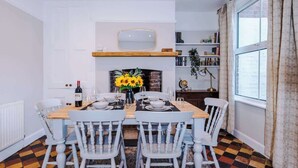 A dining area with a wooden table set for four, white chairs, a vase of sunflowers, and a bookshelf in the background.