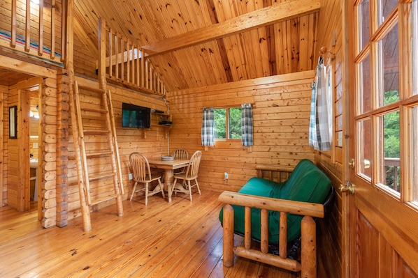 L’intérieur d’une cabane en rondins comprend un coin repas, un canapé vert, un escalier en bois et une fenêtre ornée de rideaux à carreaux.