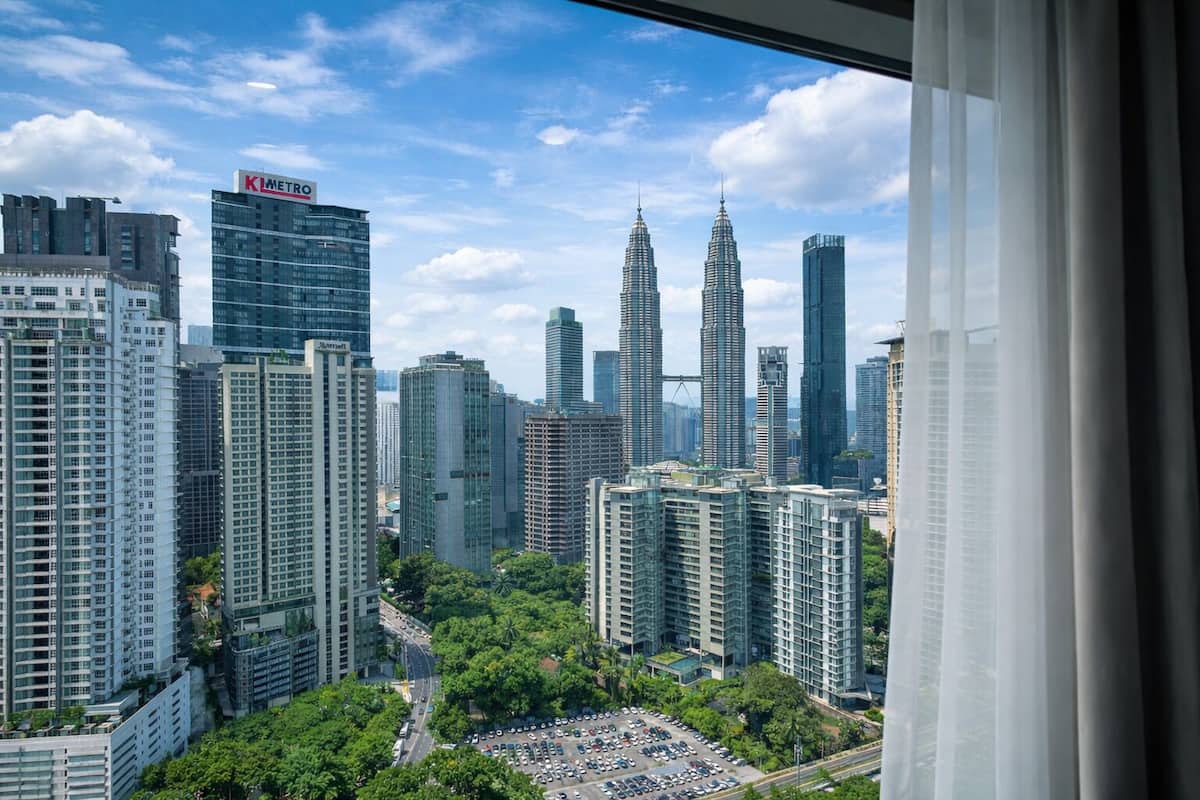A cityscape with modern high-rise buildings, including the Petronas Towers, and a view of a parking area with trees.