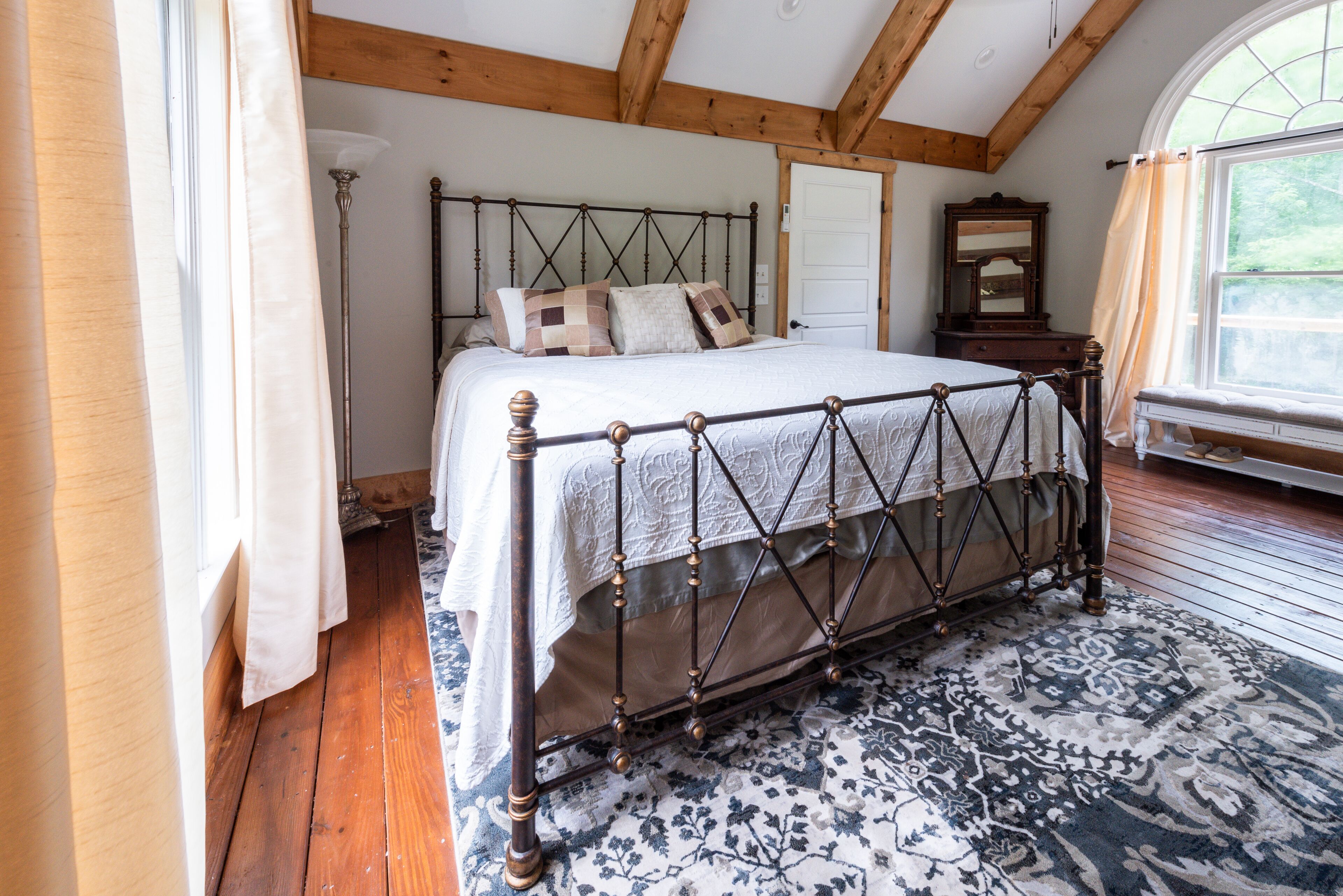 A bedroom with a large bed, wooden beams, a window with a view, and a patterned rug.