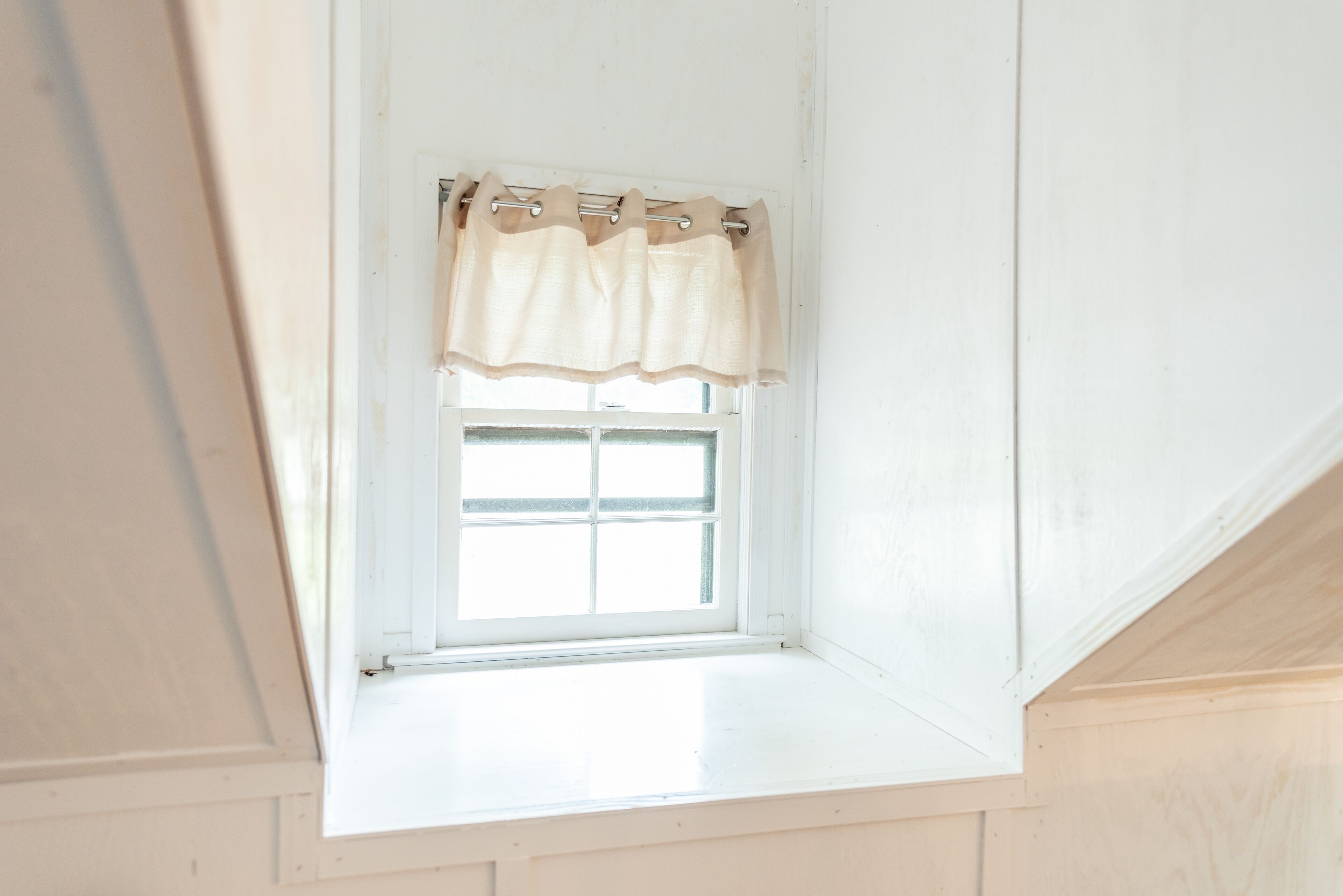 Una pequeña ventana de tejado con exterior blanco y cortina color beige.