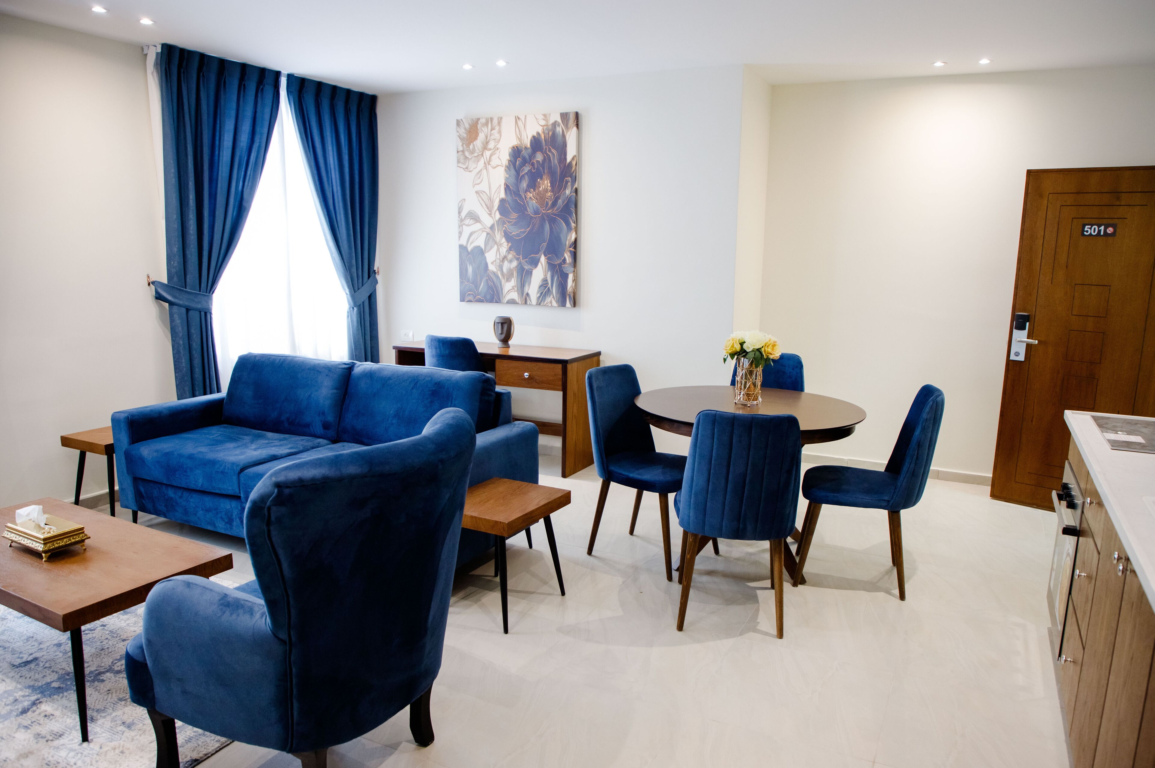 A modern living room with a blue sofa, matching chairs, a wooden coffee table, and a dining table with chairs. There is a painting on the wall and a door labeled "501".
