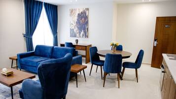 A modern living room with a blue sofa, matching chairs, a wooden coffee table, and a dining table with chairs. There is a painting on the wall and a door labeled "501".