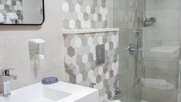 Un baño moderno con lavamanos, espejo y retrete blancos, cabina de ducha de vidrio y pared con patrón hexagonal.