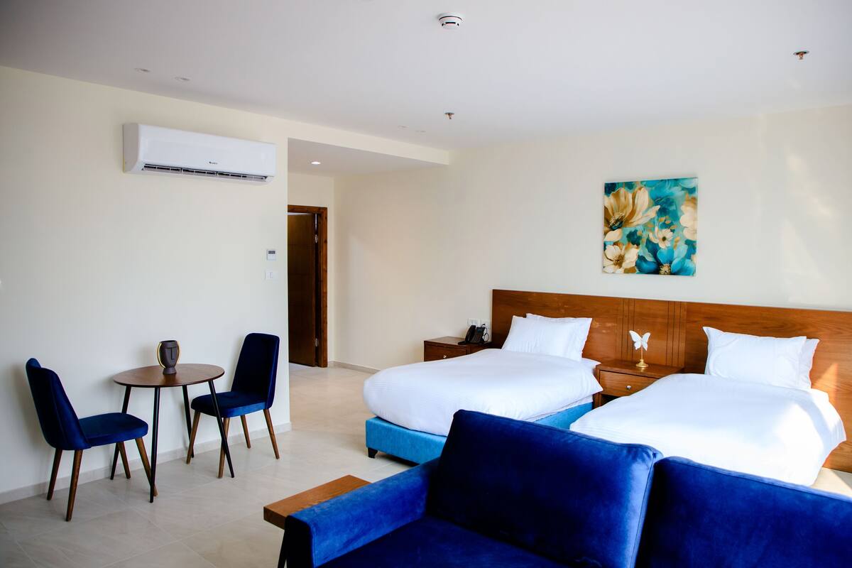 A hotel room with a large bed, two bedside tables, a small table with chairs, and an air conditioning unit on the wall.