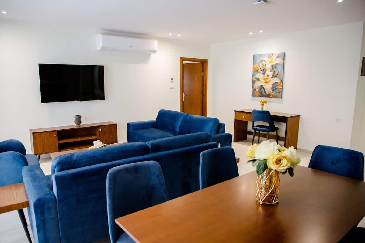 A modern room with a wooden dining table, blue sofas, a TV mounted on the wall, and a painting above the door.