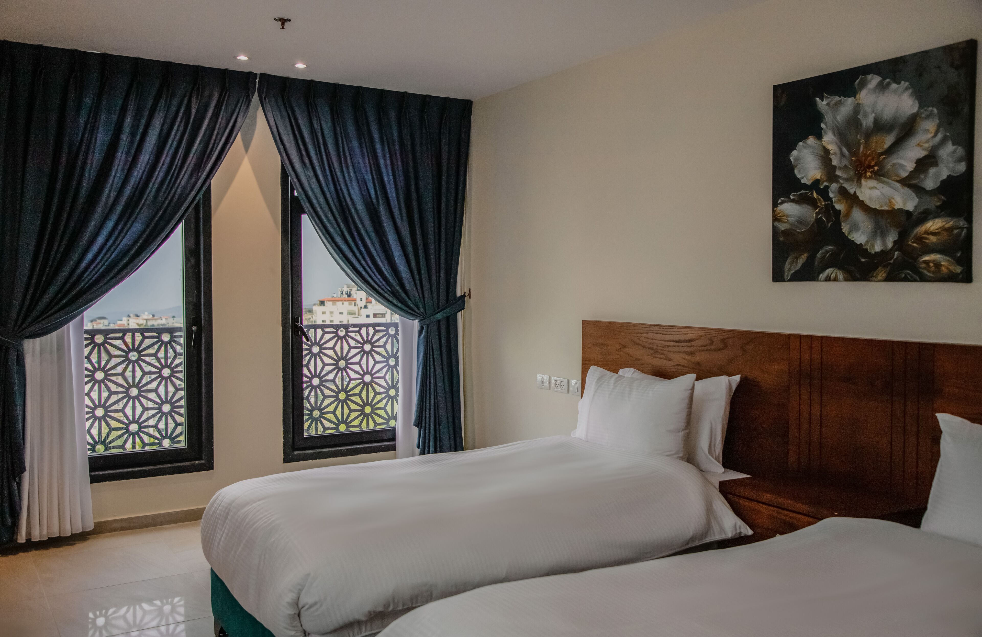 A hotel room with two beds, a large window with patterned curtains, and a painting of flowers on the wall.