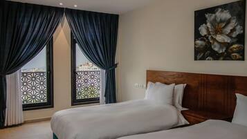 A hotel room with two beds, a large window with patterned curtains, and a painting of flowers on the wall.