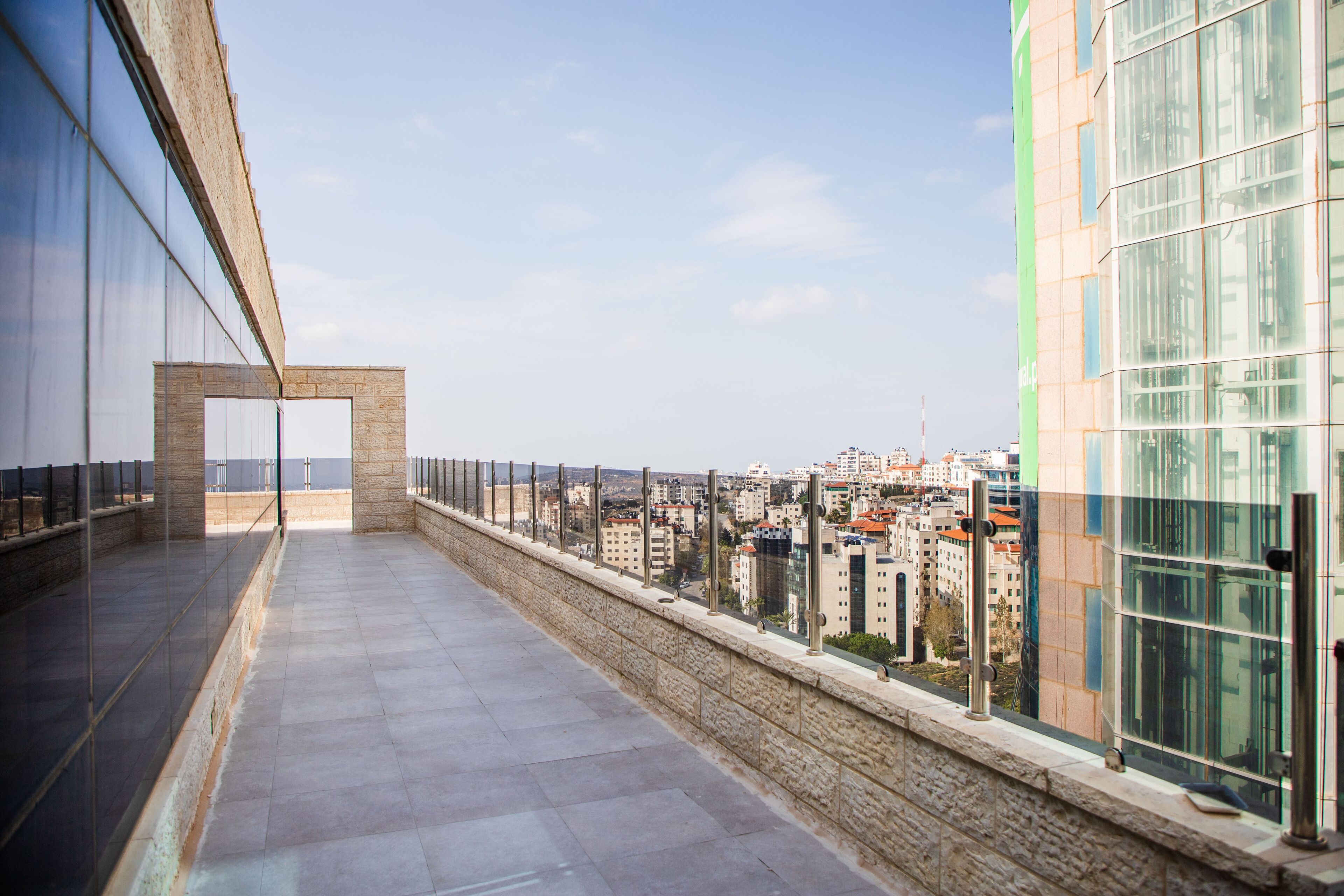 Un balcón largo y estrecho con muro de piedra y barandilla de vidrio que da a una vista panorámica de la ciudad.