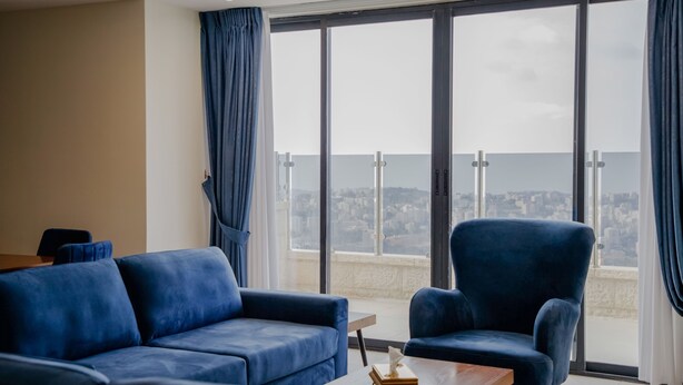 A modern living room with a blue sofa and armchair, a wooden coffee table, and large windows offering a city view. - Al-Istiklal Suites (Ramallah)