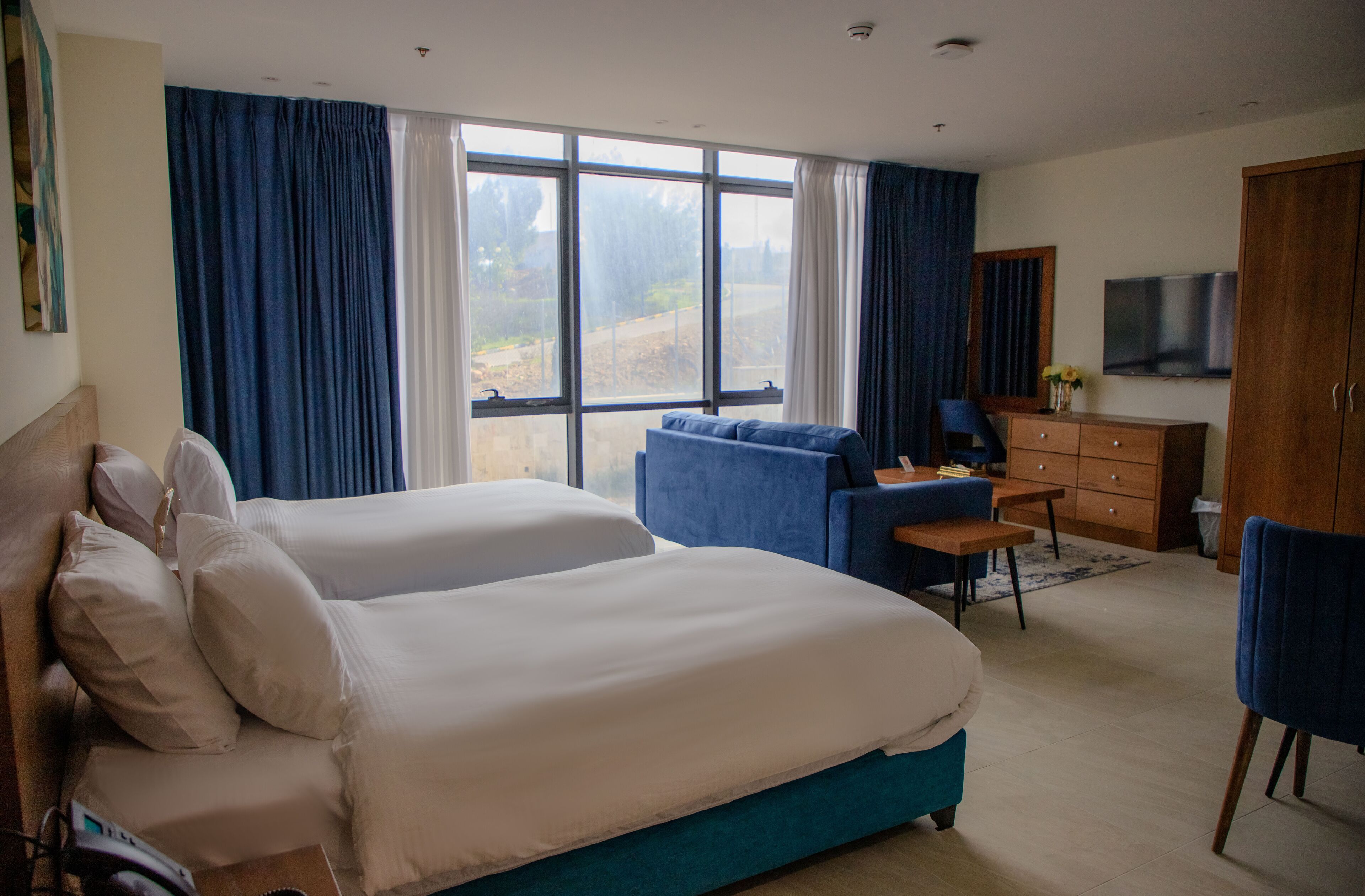 A hotel room with two beds, a desk, a chair, a TV, and large windows with curtains.