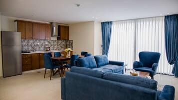A modern living room with a blue sofa set, a dining table with chairs, and a kitchen area with wooden cabinets and a stainless steel refrigerator.