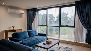 A modern living room with a large window, a blue sofa set, a wooden coffee table, and a small side table with a vase. - Al-Istiklal Suites (Ramallah)