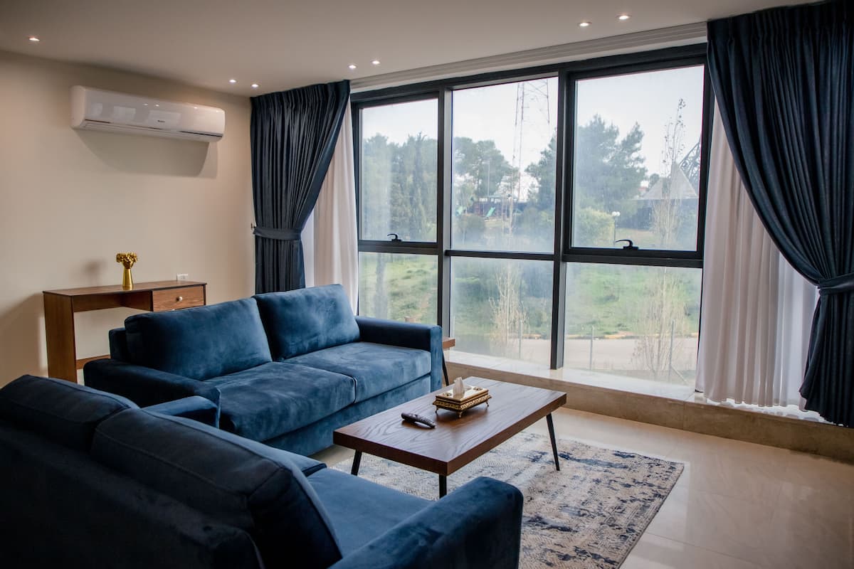 A modern living room with a large window, a blue sofa set, a wooden coffee table, and a small side table with a vase.