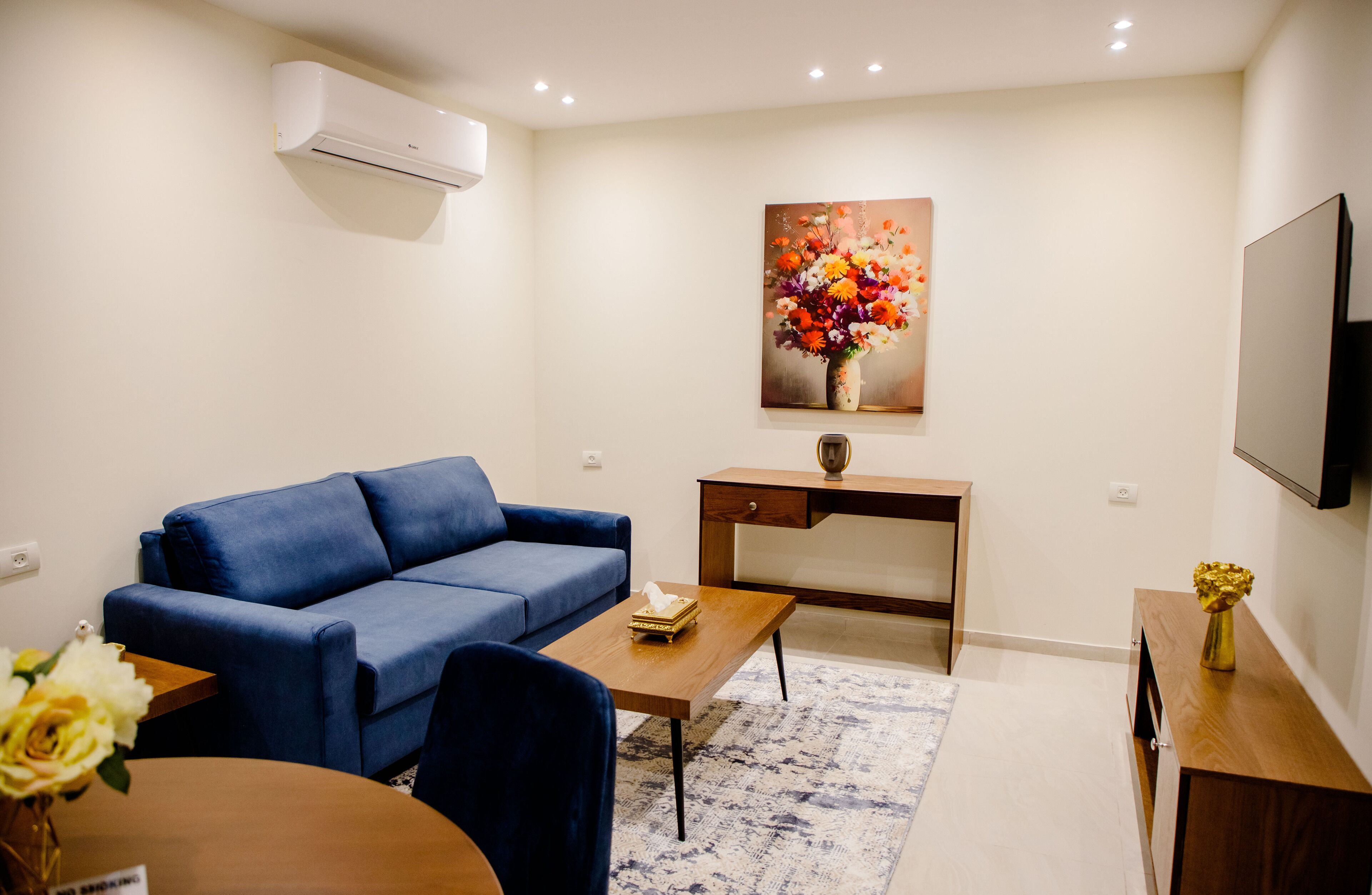A modern living room with a blue sofa, a wooden coffee table, a desk, a flat-screen TV, and a painting of flowers on the wall.