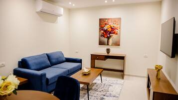 A modern living room with a blue sofa, a wooden coffee table, a desk, a flat-screen TV, and a painting of flowers on the wall.