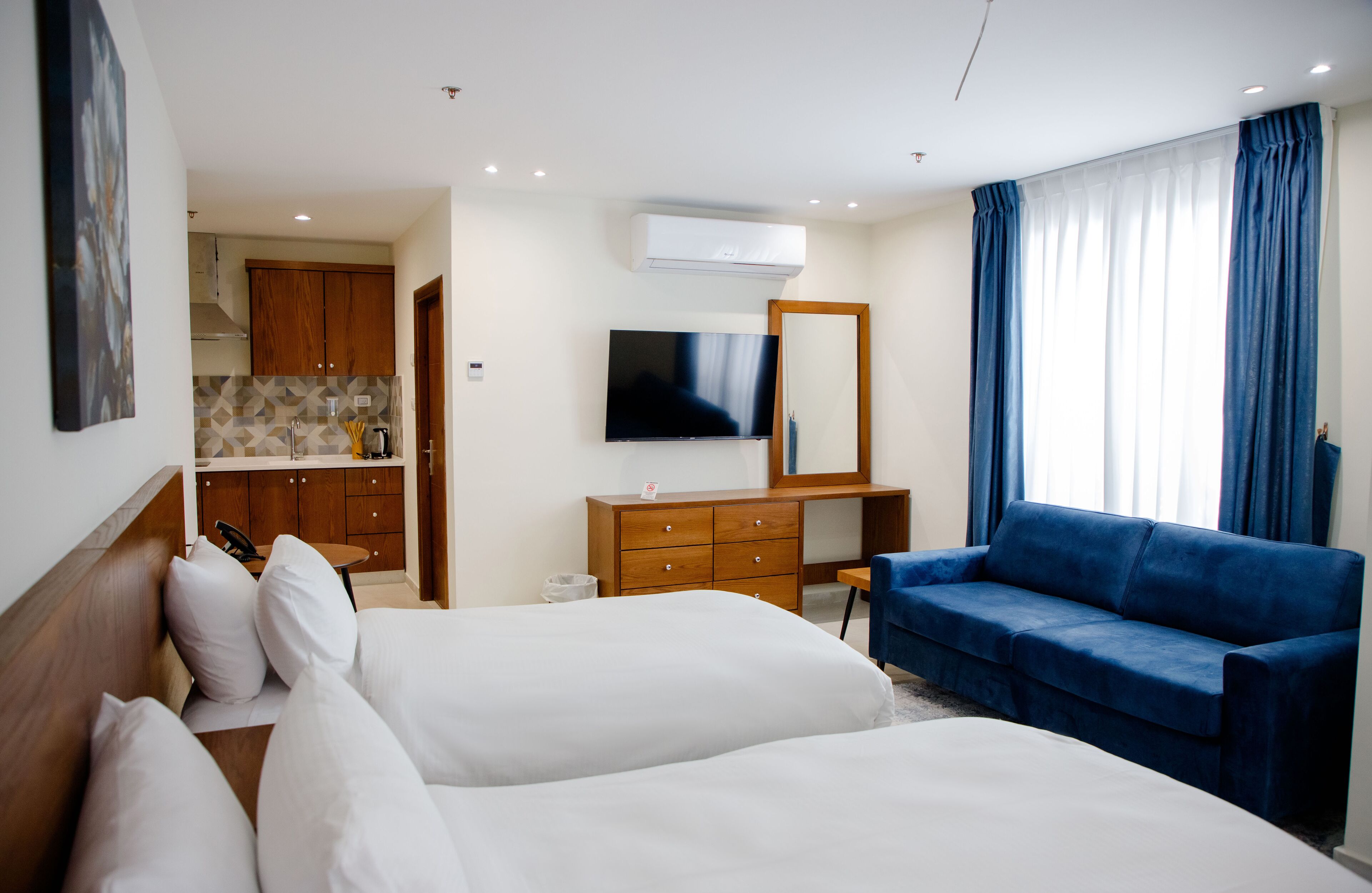 Quarto de hotel com duas camas, uma televisão, uma cômoda de madeira, um sofá azul e uma área de cozinha visível através de uma porta aberta.