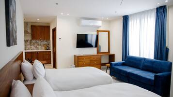 Quarto de hotel com duas camas, uma televisão, uma cômoda de madeira, um sofá azul e uma área de cozinha visível através de uma porta aberta.