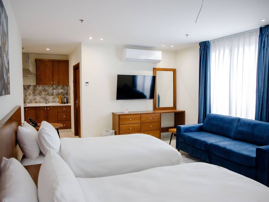 Quarto de hotel com duas camas, uma televisão, uma cômoda de madeira, um sofá azul e uma área de cozinha visível através de uma porta aberta.