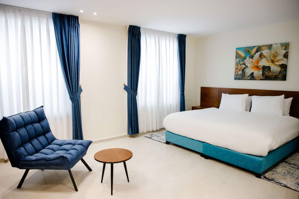 A hotel room with a large bed, a blue upholstered chair, a small round table, and a painting on the wall.