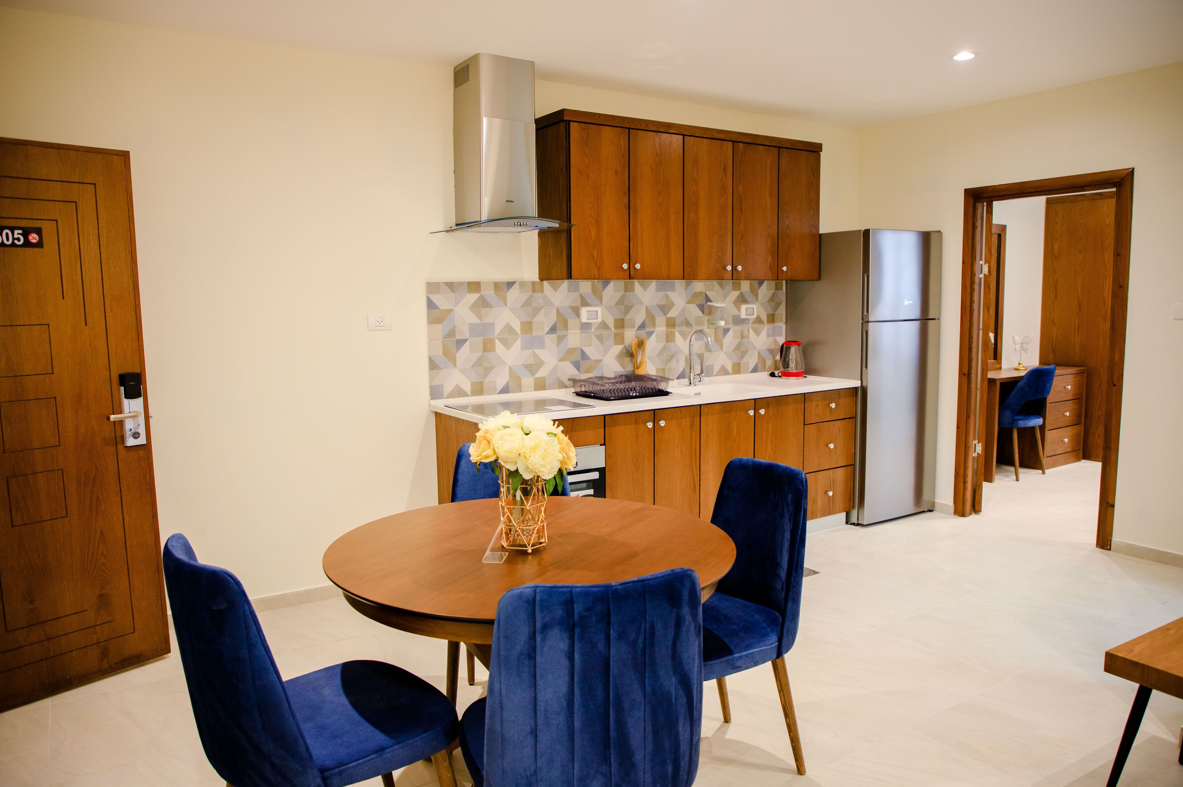 A modern kitchen with wooden cabinets, a built-in stove, and a refrigerator. There is a dining table with blue chairs and a vase of flowers.