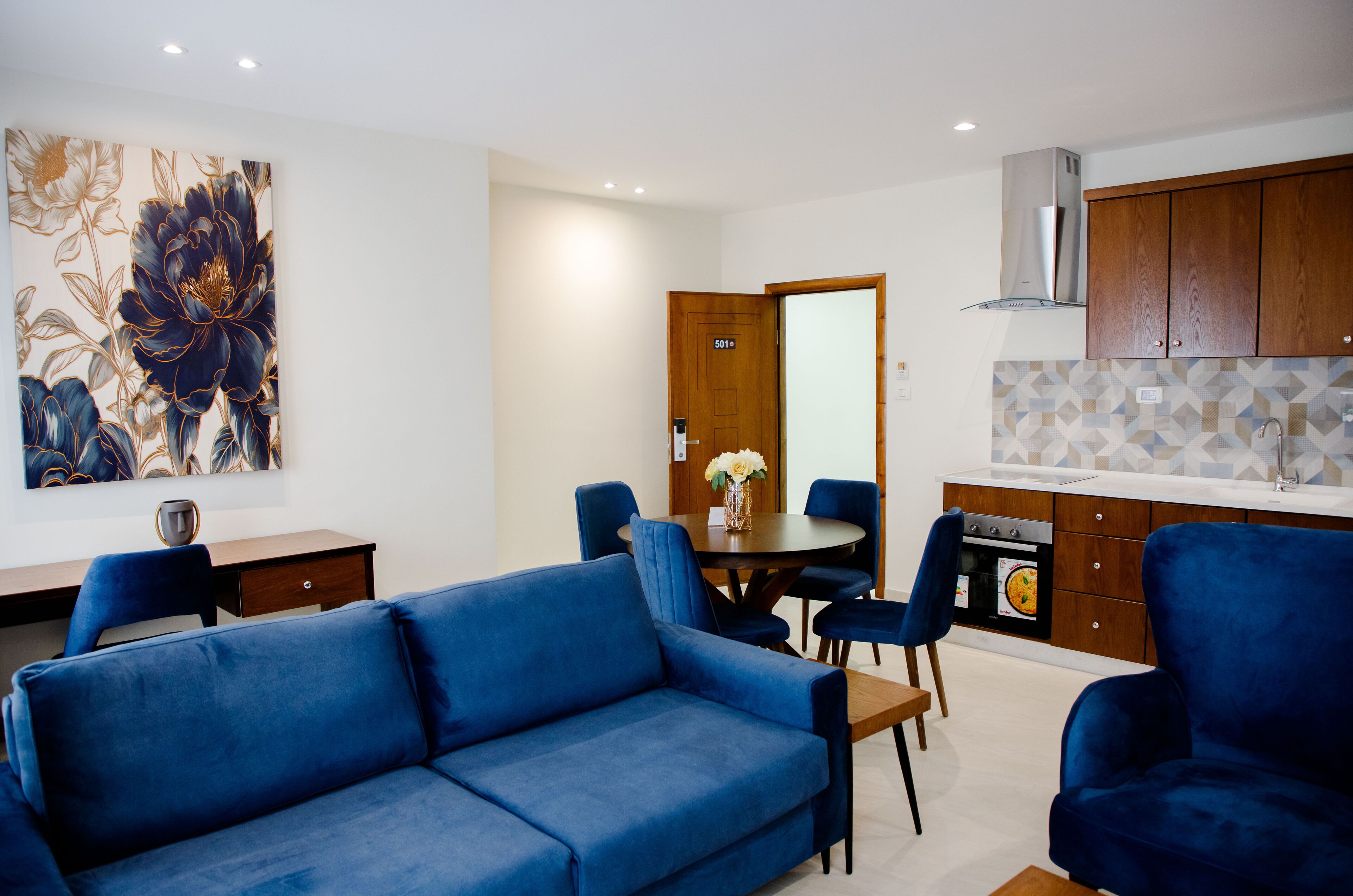 Un salon moderne avec un canapé bleu, une table à manger avec des chaises et un coin cuisine doté de meubles en bois et d’un dosseret carrelé.
