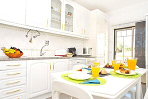 A bright kitchen with white cabinets, a microwave, and a dining table set with plates, cutlery, and a fruit bowl.