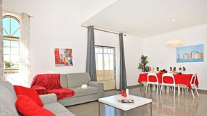 A modern living room with a grey sofa, red cushions, a white coffee table, and a dining area with a red tablecloth. - Cozy Apartment w/ Pool in Albufeira (Albufeira)