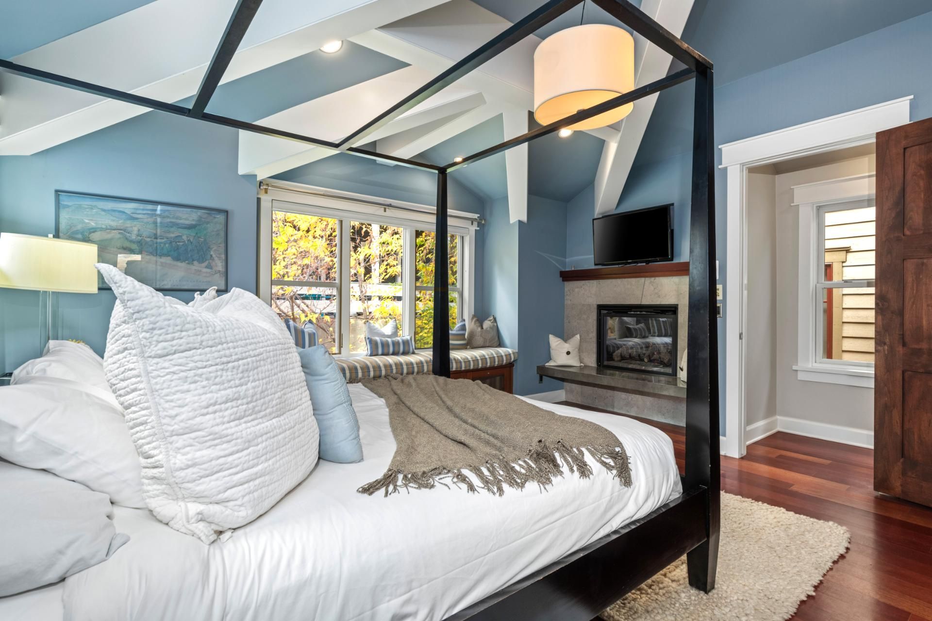 Un dormitorio con cama con dosel, chimenea, televisor y ventana con vistas a árboles.