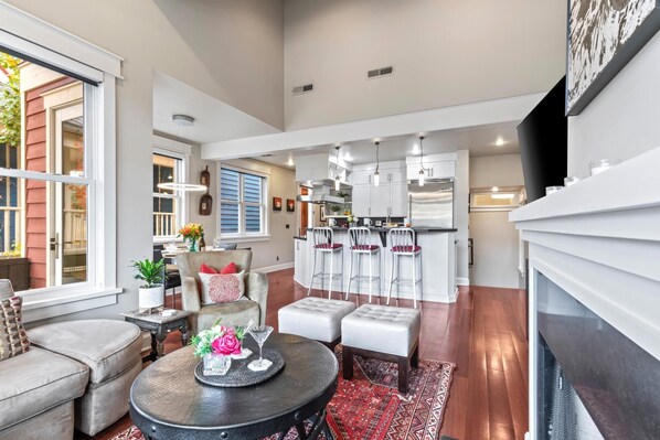 A spacious living room with a fireplace, a dining area with a kitchen in the background, and a large window providing natural light.