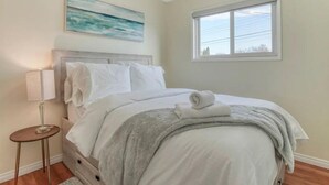A neatly made bed with white linens and a grey blanket, a bedside lamp, and a painting of a seascape above it. - Fort Charm (Fort Saskatchewan)
