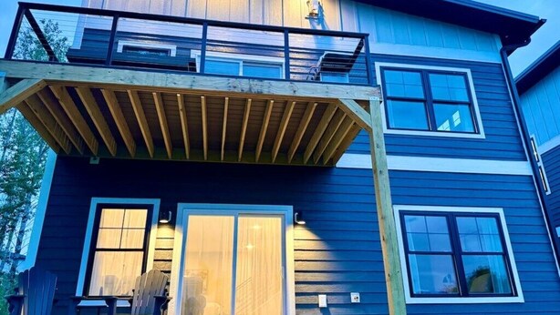 Een tweelaags huis met een houten terras, grote schuifdeuren en een zichtbare trap.