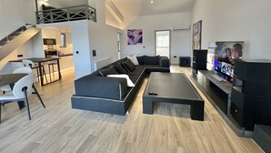 Un salon moderne avec un canapé d’angle, une table basse et un téléviseur. On aperçoit à l’arrière-plan un coin cuisine et un escalier menant à un étage supérieur.