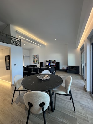A modern living room with a round dining table, comfortable seating, a flat-screen TV, and a fireplace.