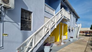 Un edificio con una escalera blanca que conduce a una puerta amarilla, unidades de aire acondicionado y plantas en macetas.