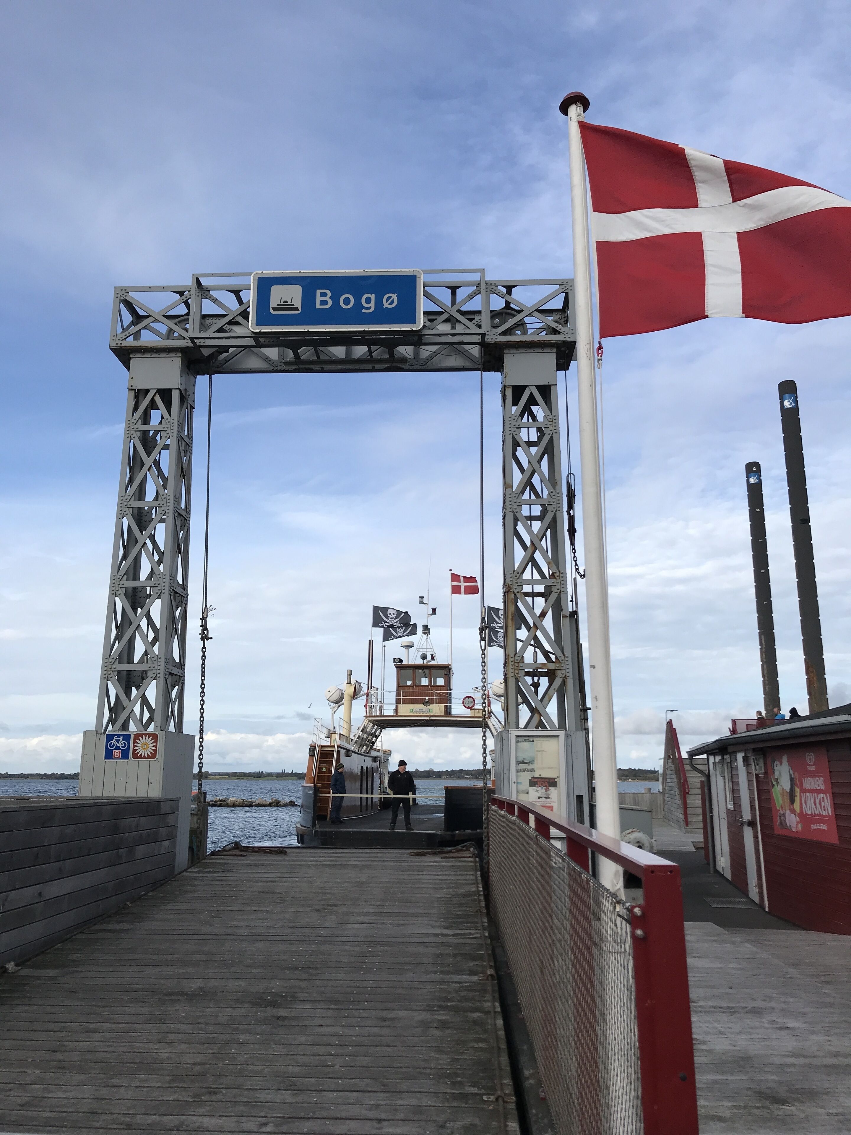 Entrada de un muelle con un letrero que dice "Boga", una bandera y un pequeño edificio con un letrero.