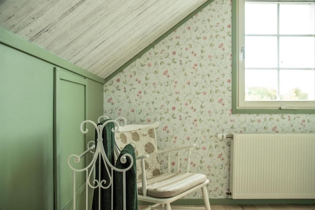 Une pièce avec un plafond en pente, un radiateur, un papier peint à motifs floraux et une fenêtre.