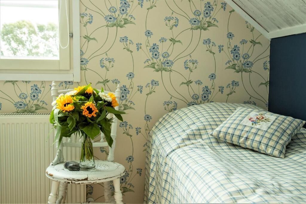 Una camera da letto con carta da parati a motivi floreali, un letto con copriletto a quadretti e un tavolino con un vaso di fiori.