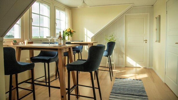 Un comedor con mesa de madera y sillas negras, una lámpara colgante y vistas al exterior a través de grandes ventanales.