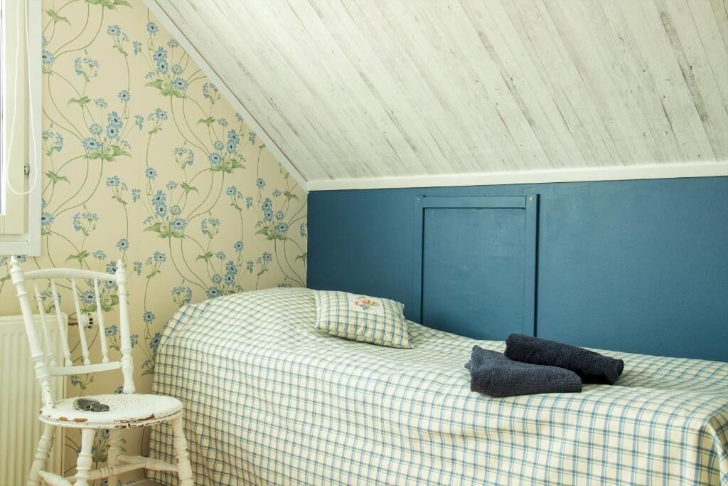 A bedroom with a checkered bedspread, a blue headboard, a white chair, and floral wallpaper.