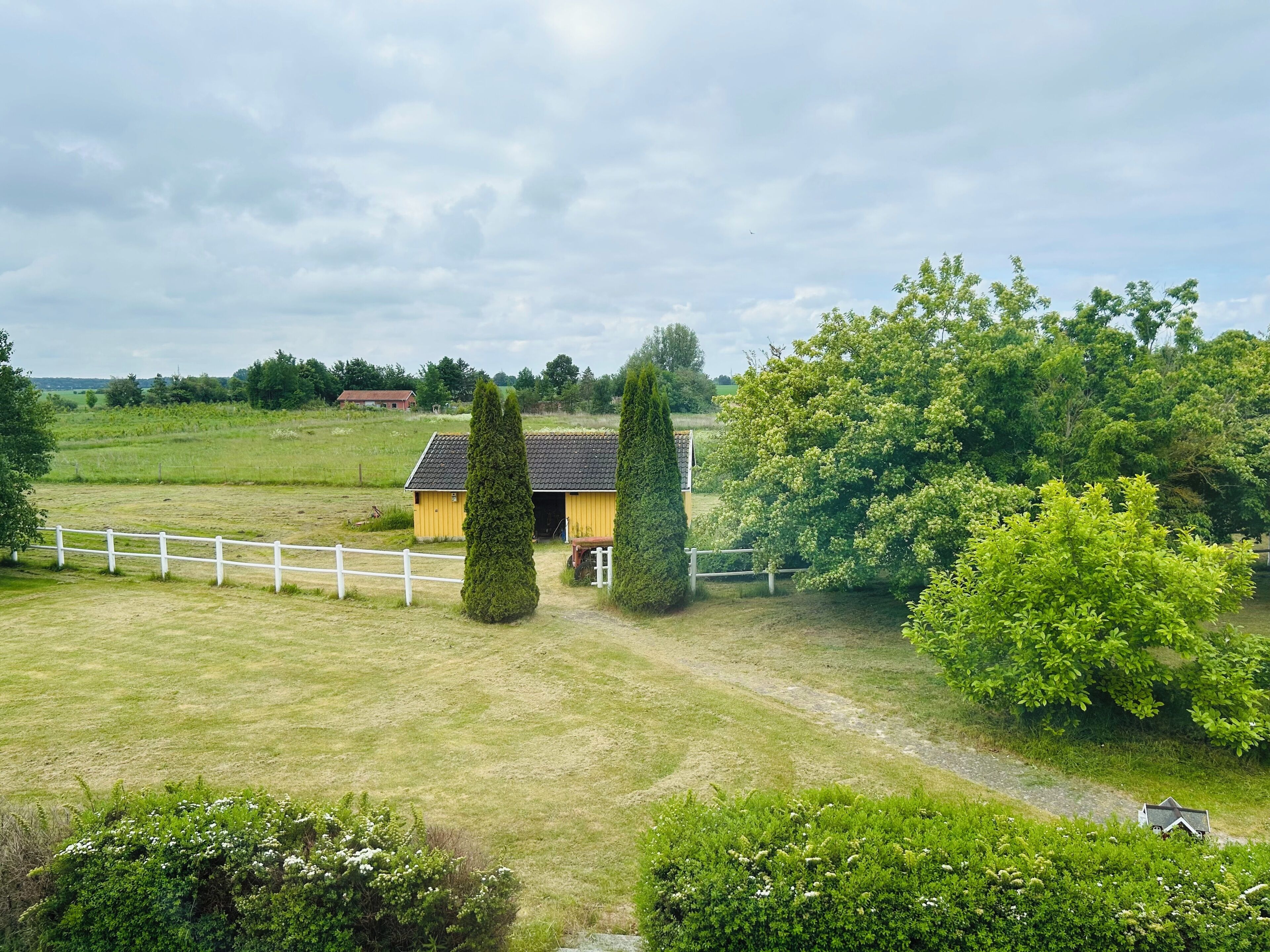 A rural landscape with a yellow building, white fence, and green trees.