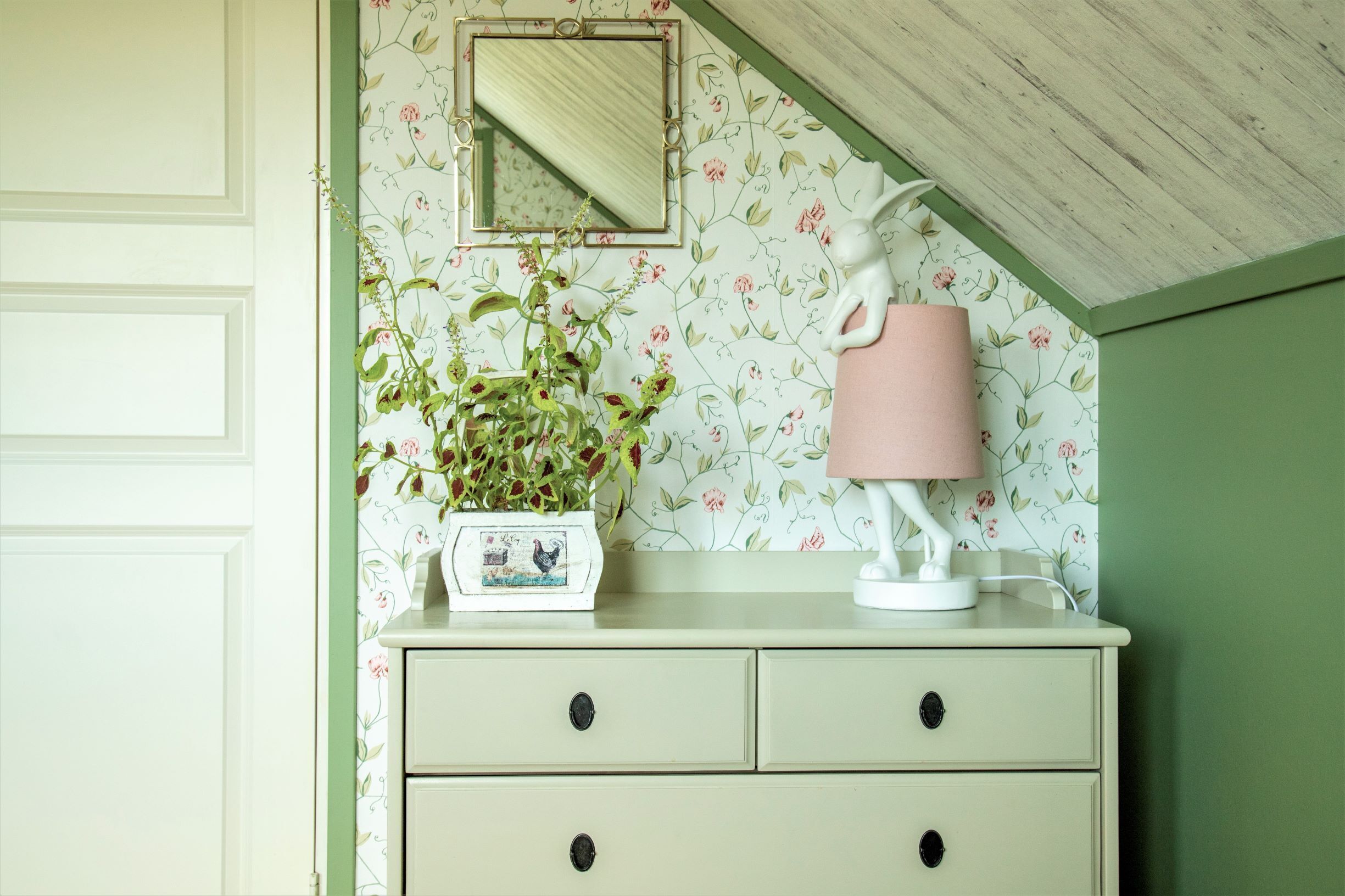 A room with a green wall, a white dresser with two drawers, a mirror, a pink lamp, and a potted plant.