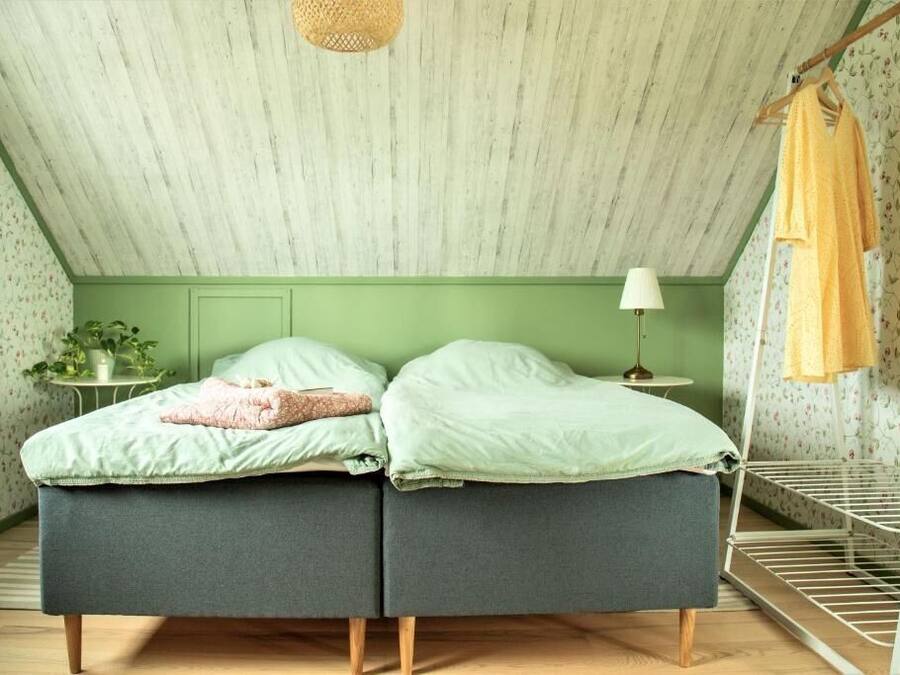 Una habitación con techo inclinado, dos camas con ropa de cama verde, una mesita con una lámpara y un sillón colgante.