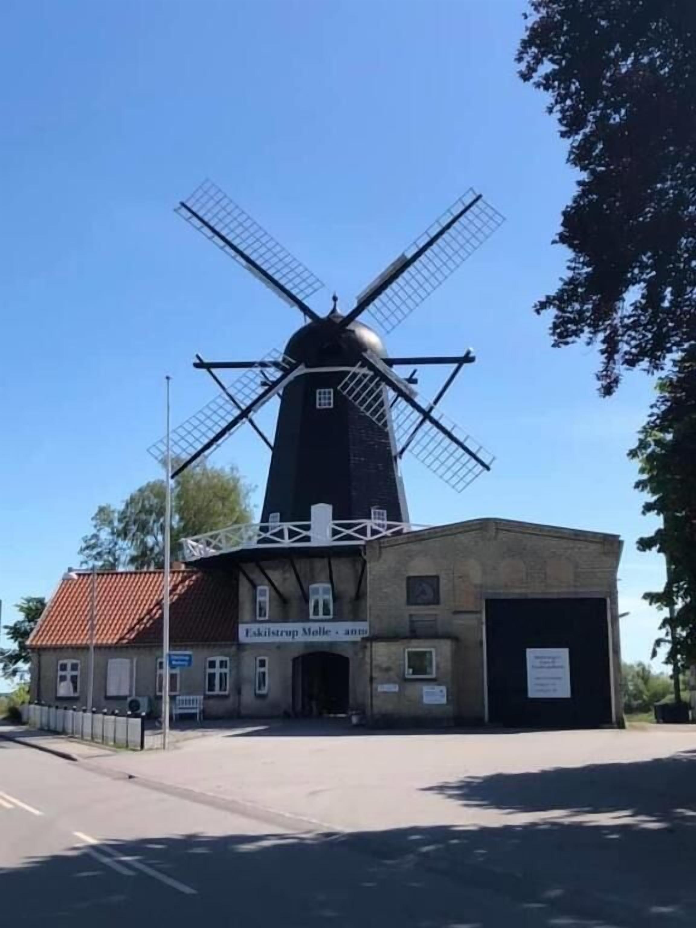 Un molino de viento tradicional con un letrero que dice "Fiskilstrup Mølle" frente a un edificio con techo de tejas y una señal.