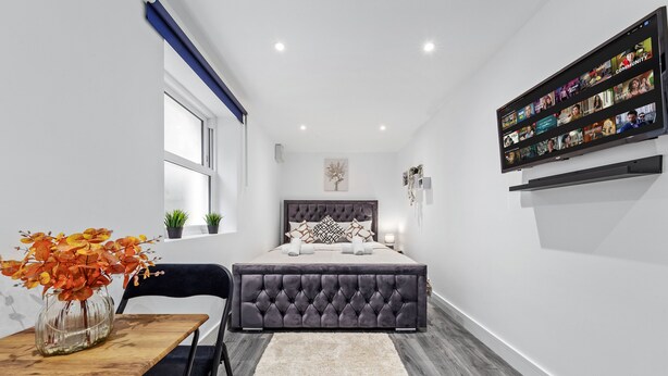 A modern bedroom with a large bed, a TV mounted on the wall, a small table with a vase of flowers, and a window with a view of the outside. - Luxury Ensuite Room in Finsbury Pak London N19 (London)