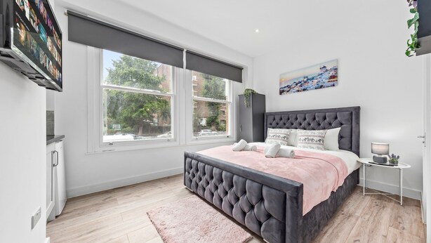A modern bedroom with a large bed, a TV mounted on the wall, and a window with a view of the outside. - Charming Ensuite Room in Finsbury Park London N19 (London)