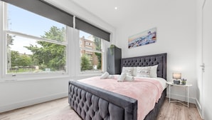 A bedroom with a large window, a bed with a tufted headboard, a bedside table with a lamp, and a painting on the wall. - Charming Ensuite Room in Finsbury Park London N19 (London)