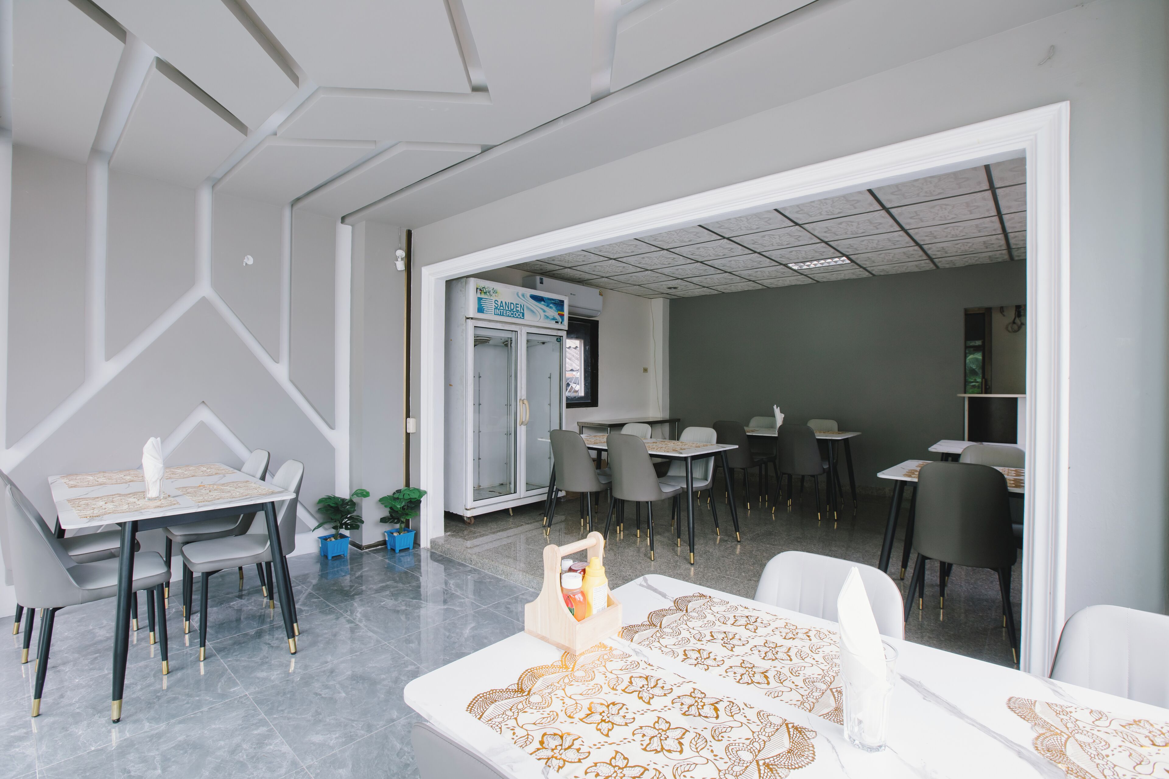 A modern dining area with white tables and chairs, a visible sandwich shop sign, and a clean, geometric ceiling design.
