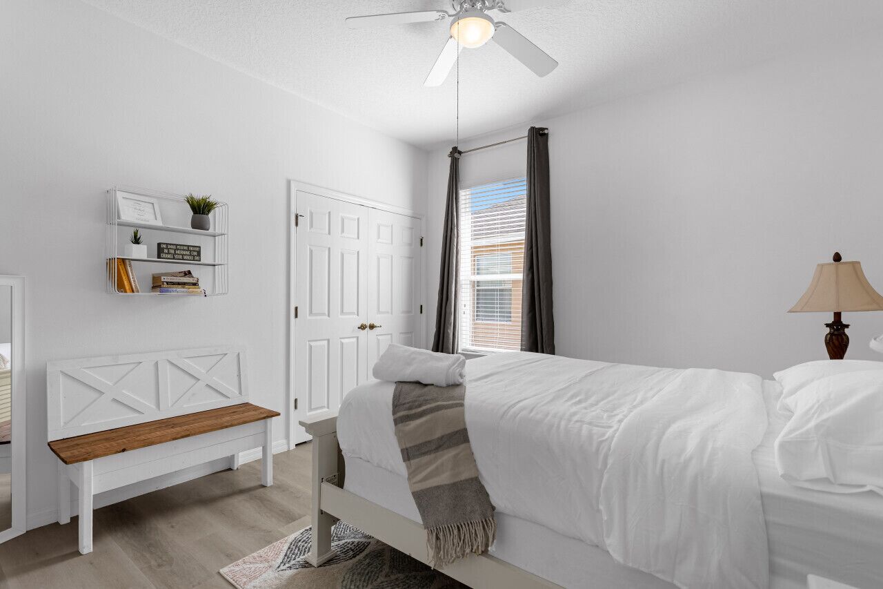 Ein Schlafzimmer mit einem Bett, einer weißen Bank, einem Regal mit Büchern und einer Pflanze, einem Deckenventilator und einem Fenster mit Vorhängen.