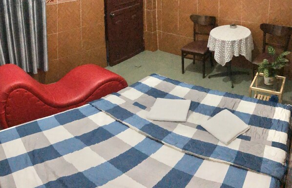 A room with a checkered bedspread, a red chair, a small round table with a white tablecloth, and a potted plant.